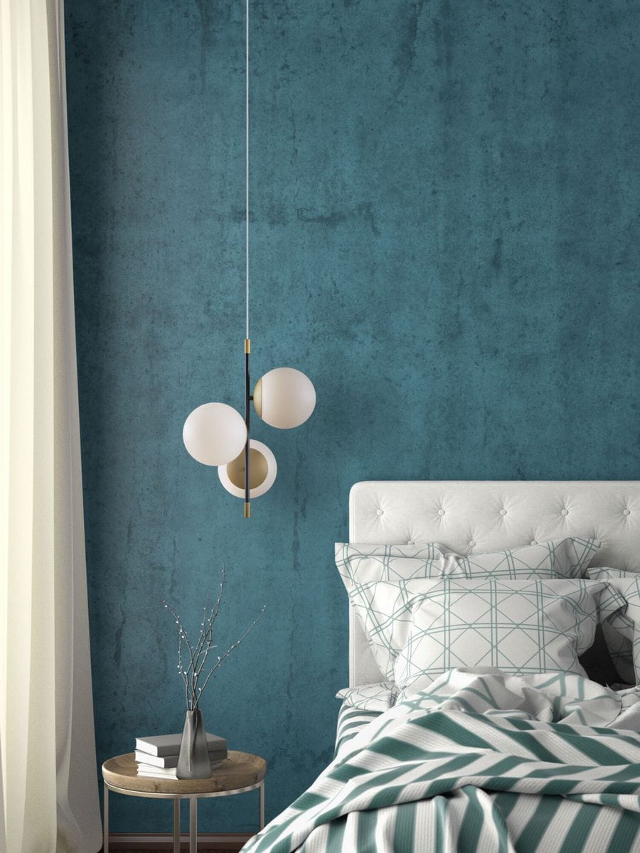 Lampa wisząca Nostalgia 3-punktowa w kolorze złotym, z trzema okrągłymi, białymi kloszami. Zawieszona na cienkim przewodzie, umieszczona nad łóżkiem w nowoczesnej sypialni z niebieską ścianą i drewnianym stolikiem nocnym.