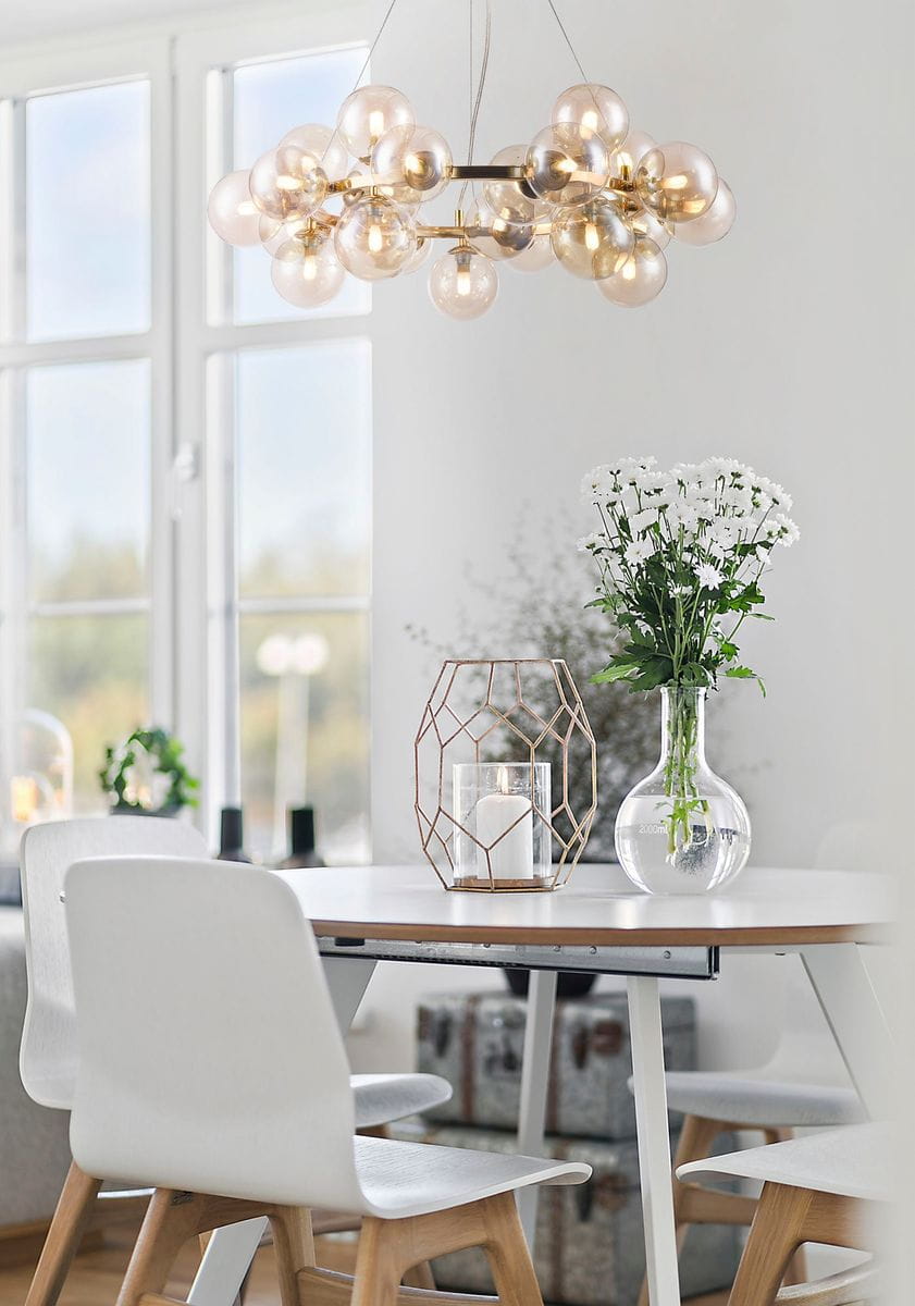 Lampa wisząca Dallas 25-punktowa w kolorze złotym, z okrągłymi kloszami w odcieniach przezroczystych. Zawieszona na cienkich linkach, umieszczona nad stołem w jasnym wnętrzu. W tle widoczne białe krzesła i dekoracje: wazon z kwiatami oraz świecznik.