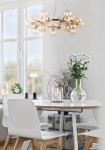 Lampa wisząca Dallas 25-punktowa w kolorze złotym, z okrągłymi kloszami w odcieniach przezroczystych. Zawieszona na cienkich linkach, umieszczona nad stołem w jasnym wnętrzu. W tle widoczne białe krzesła i dekoracje: wazon z kwiatami oraz świecznik.
