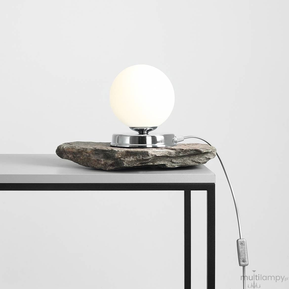 Lampa stołowa Ball D14 z chromowanym podstawą i białą, okrągłą kulą, umieszczona na naturalnym kamieniu. Kabel z włącznikiem wychodzi z podstawy. Ujęcie frontalne na stole o prostokątnej konstrukcji.