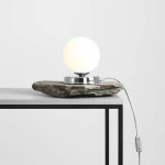 Lampa stołowa Ball D14 z chromowanym podstawą i białą, okrągłą kulą, umieszczona na naturalnym kamieniu. Kabel z włącznikiem wychodzi z podstawy. Ujęcie frontalne na stole o prostokątnej konstrukcji.