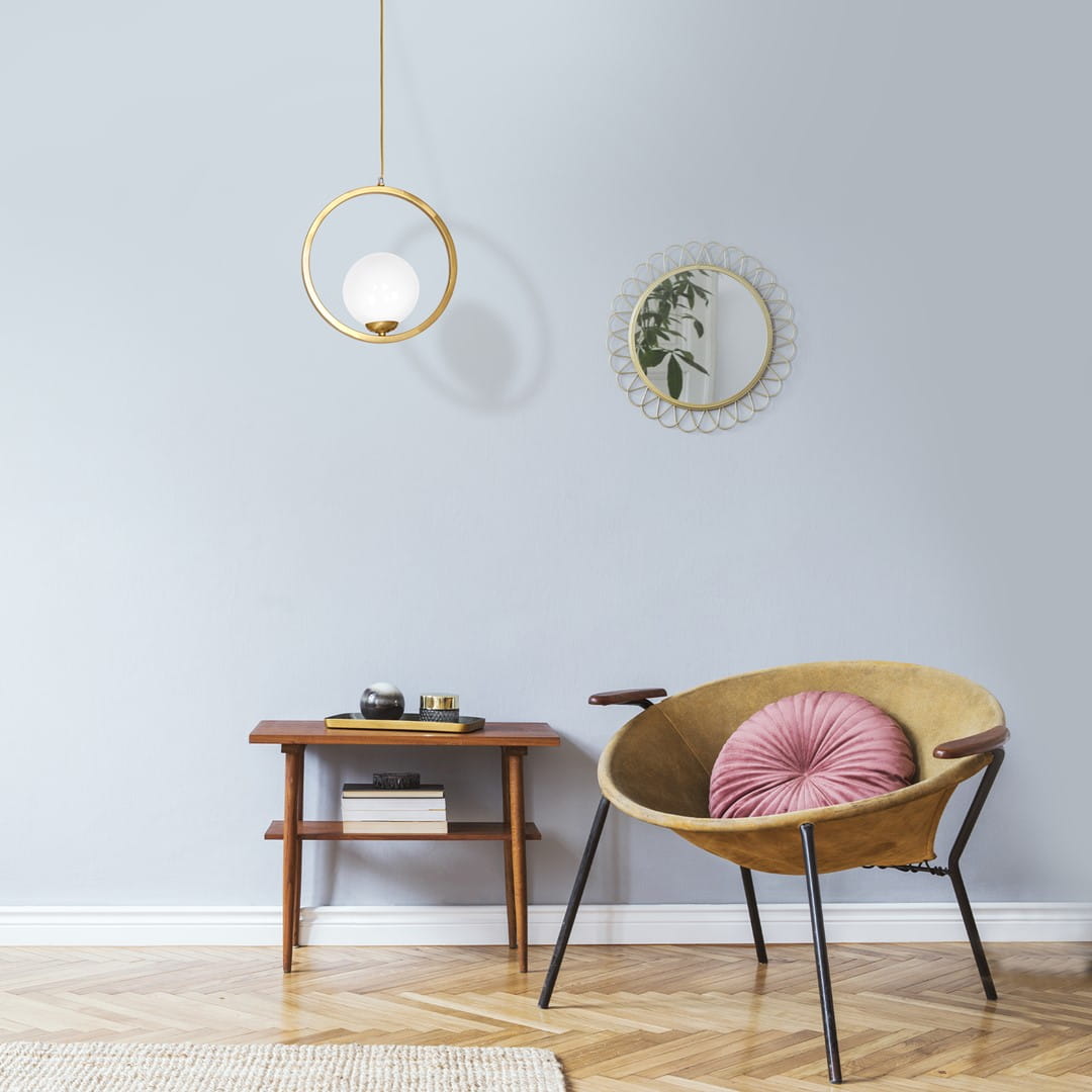 Lampa wisząca Vienna 1-punktowa MLP5573 w złotym okręgu z białym kloszem, zawieszona nad drewnianym stolikiem. Tło w pastelowym odcieniu, w aranżacji z krzesłem i lustrem, tworzy nowoczesny, minimalistyczny wygląd.