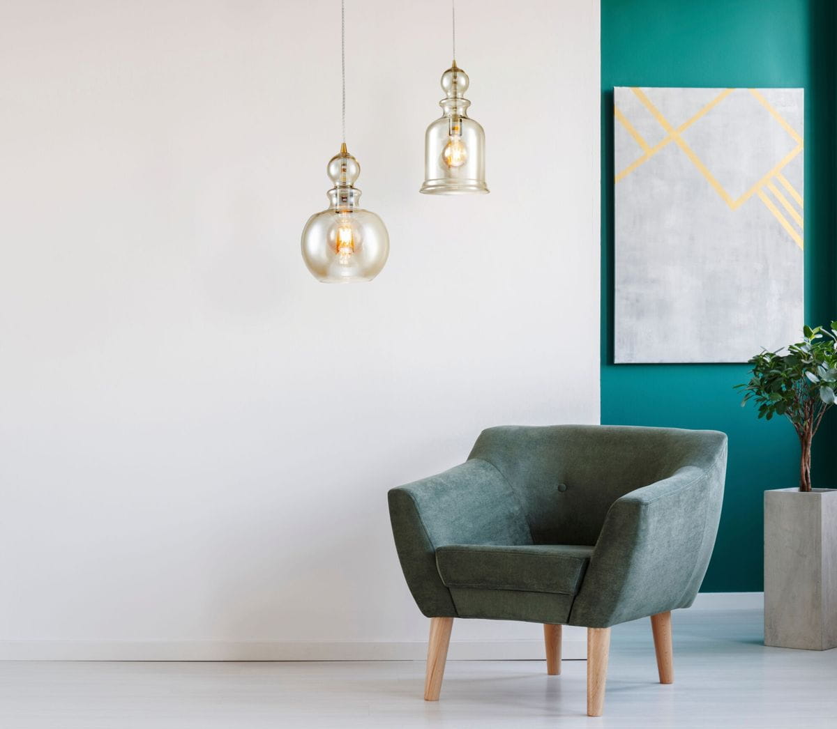 Brązowa lampa wisząca 1-punktowa Tone P003PL-01BZ w kształcie kulistym, wykonana z przezroczystego szkła, z widoczną żarówką. Zawieszona na cienkim kablu, w otoczeniu zielonej ściany i nowoczesnego fotela.