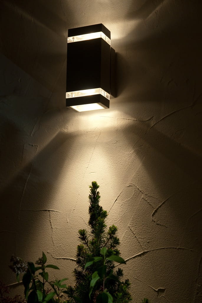 Ciemnoszary kinkiet elewacyjny Focus LED 2-punktowy, zamontowany na ścianie, emitujący światło w górę i w dół. W tle widoczna roślina. Lampa ma prostokątny kształt, z przezroczystymi elementami u góry i na dole, tworzącymi efekt świetlny.