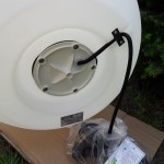 Lampa ogrodowa Cumulus M w kolorze białym, z oprawą na żarówkę E27. Widoczny tył lampy z kablem oraz miejscem na mocowanie. Produkt leży na kartonie, w tle zieleń roślinności.