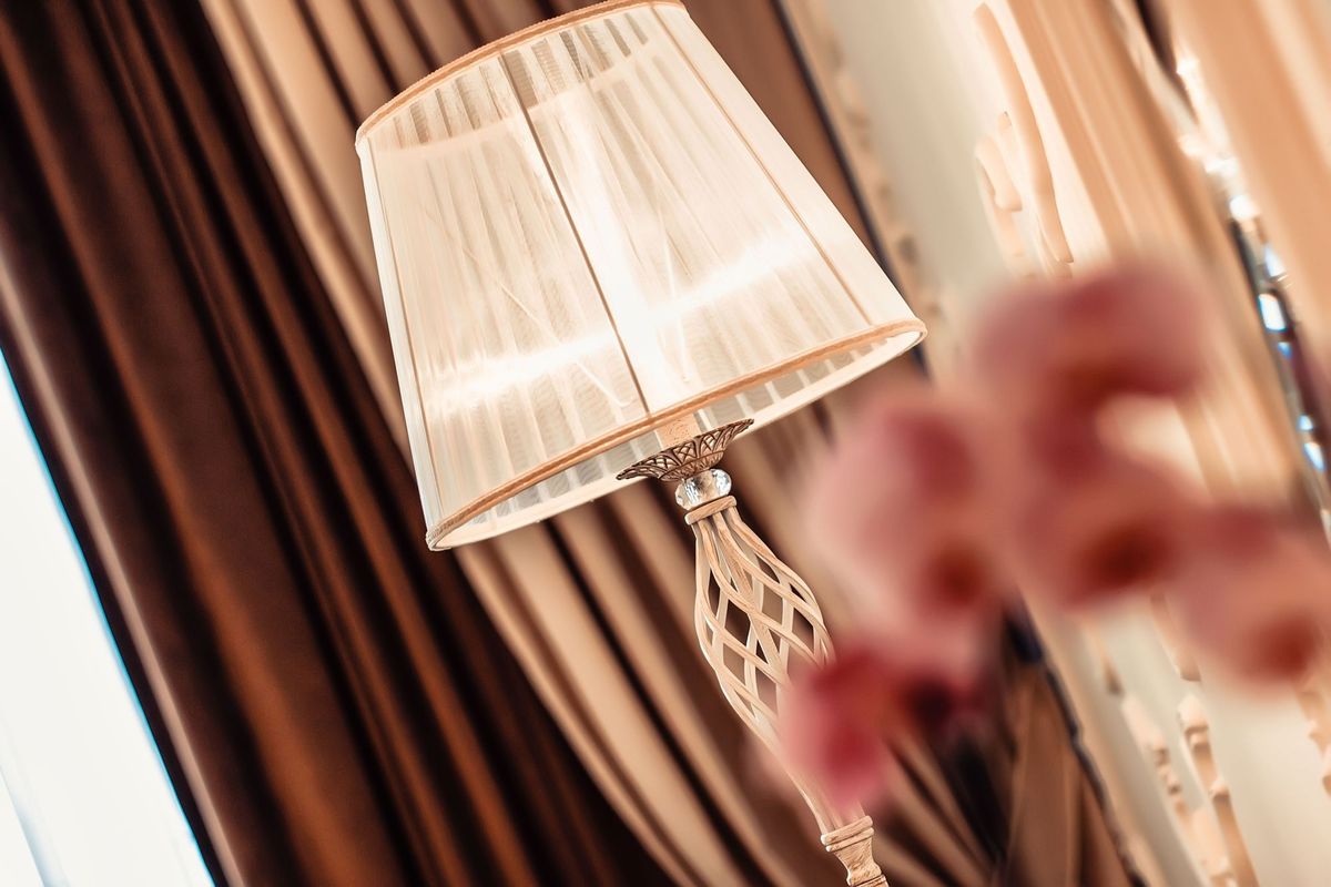 Lampa podłogowa Grace, 1-punktowa, w kolorze antycznej bieli. Stojąca, z cylindrycznym kloszem i ozdobnym, plecionym trzonem. Widoczna w eleganckim wnętrzu, z tłem ciemnych zasłon. Klosz emitujący ciepłe światło, detal z kryształową dekoracją.