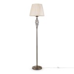 Lampa podłogowa Grace w kolorze patynowym, z okrągłą podstawą i smukłym, dekoracyjnym trzonem. Górna część z plisowanym kloszem w odcieniu jasnego beżu, stojąca na białym tle. Kabel z włącznikiem widoczny obok.