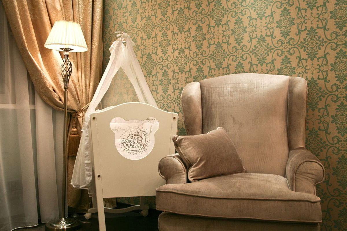 Lampa podłogowa Grace w kolorze patyny z kloszem w odcieniu jasnego beżu, stojąca obok wygodnego fotela. W tle dekoracyjna tapeta w zielone wzory oraz zasłony w odcieniu złotym. W aranżacji widoczne również dziecięce łóżeczko z przezroczystym baldachimem.