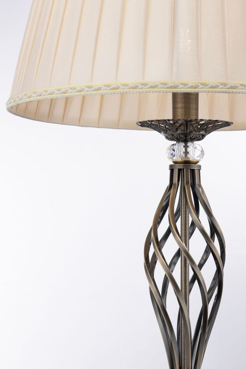 Lampa podłogowa Grace 1-punktowa w kolorze patyny z abażurem w odcieniu kremowym. Metalowa podstawa z plecionym wzorem, ozdobiona kryształowym detalem. Abażur z plisowaniem i dekoracyjną taśmą. Ujęcie z boku, ukazujące szczegóły konstrukcji.