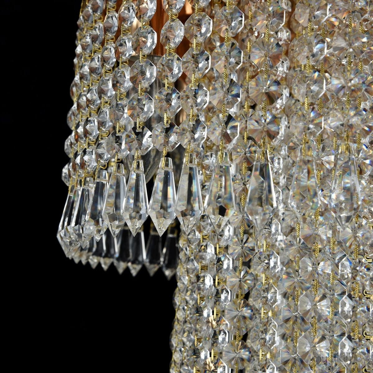 Lampa sufitowa Cascade 12-punktowa z kryształowymi zdobieniami w kształcie diamentów, zawieszona na złotych łańcuszkach. Widoczna jest tekstura kryształów, które odbijają światło, tworząc efekty świetlne. Kąt ujęcia z boku, na czarnym tle.