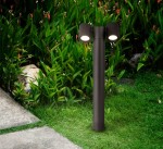 Lampa stojąca ogrodowa Wall Street H55 w kolorze czarnym, z dwiema okrągłymi głowicami oświetleniowymi, umieszczona na prostym, cylindrycznym słupku. Ujęcie z boku, na tle zielonej trawy i roślinności.