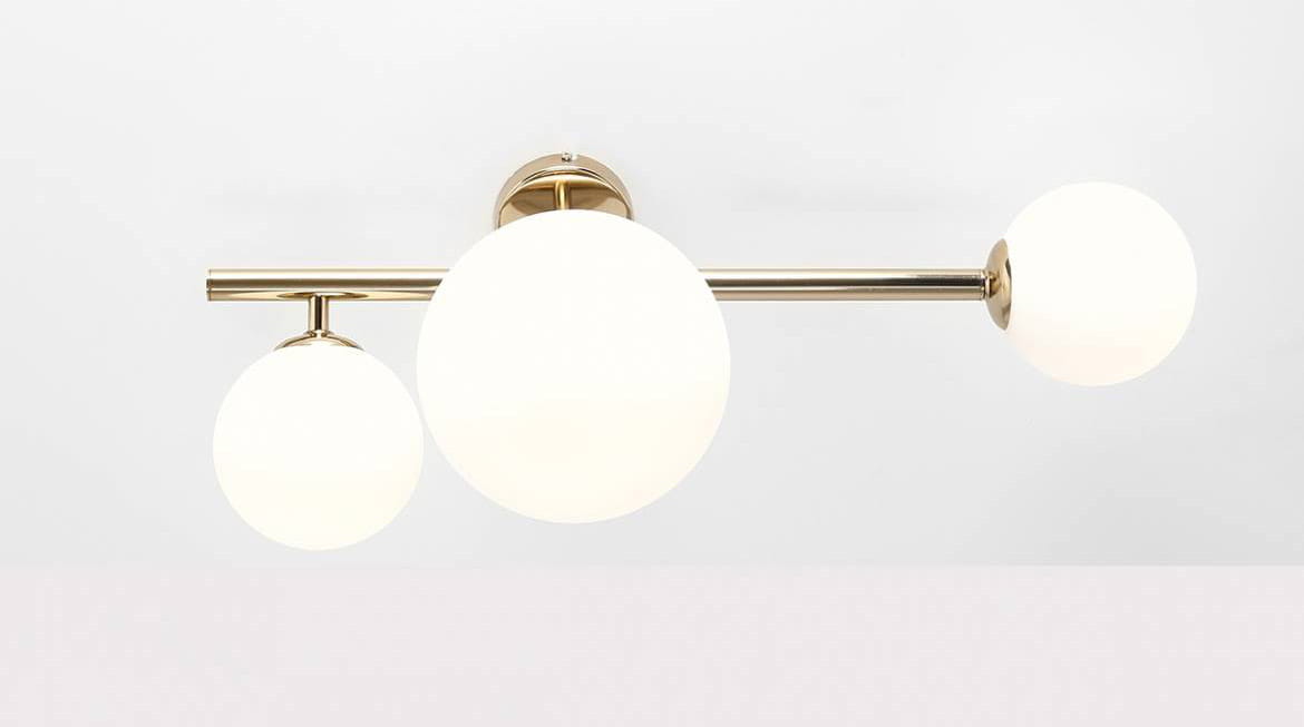 Złota lampa sufitowa Dione 3-punktowa z trzema białymi, okrągłymi kloszami w matowym wykończeniu. Ujęcie frontalne, lampa zamontowana na suficie, z prostą konstrukcją. Klosze umieszczone na różnych wysokościach, wykonane z szkła.