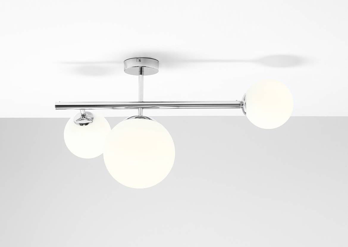 Lampa sufitowa Dione 3-punktowa w kolorze chromu z trzema białymi, okrągłymi kloszami. Ujęcie frontalne, produkt zamontowany na suficie, z widocznym chromowanym stelażem. Klosze emitują miękkie światło, tworząc nowoczesny efekt oświetleniowy.