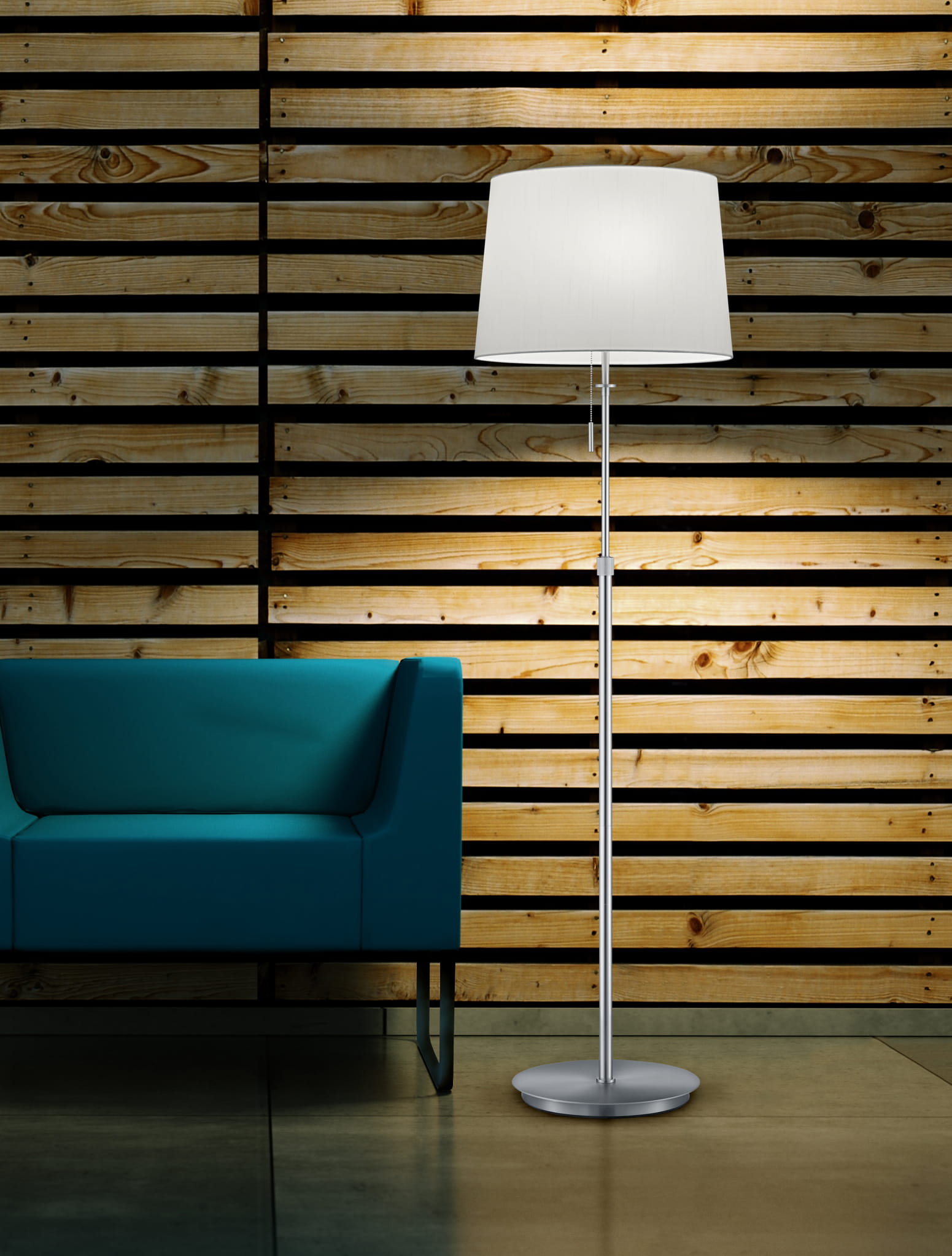 Lampa podłogowa Lyon 3-punktowa, niklowana, z białym abażurem, stojąca obok niebieskiej sofy. Ujęcie z boku, na tle drewnianej ściany. Wysoka, prosta konstrukcja z okrągłą podstawą.
