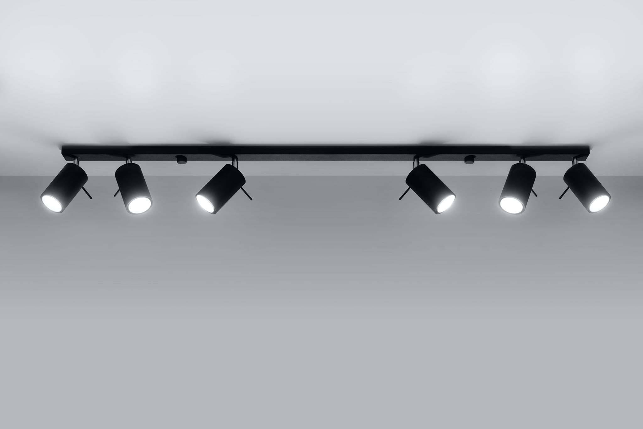 Czarna lampa sufitowa typu spot z sześcioma punktami świetlnymi, zamontowana na prostokątnej listwie. Lampy mają cylindryczny kształt i są skierowane w różne strony, co pozwala na regulację kierunku światła. Tło jasne, minimalistyczne.