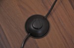 Czarny włącznik lampy podłogowej, okrągły kształt, z przyciskiem na górze. Kabel zasilający wychodzi z boku. Tło to drewniana podłoga, co podkreśla elegancki styl produktu.