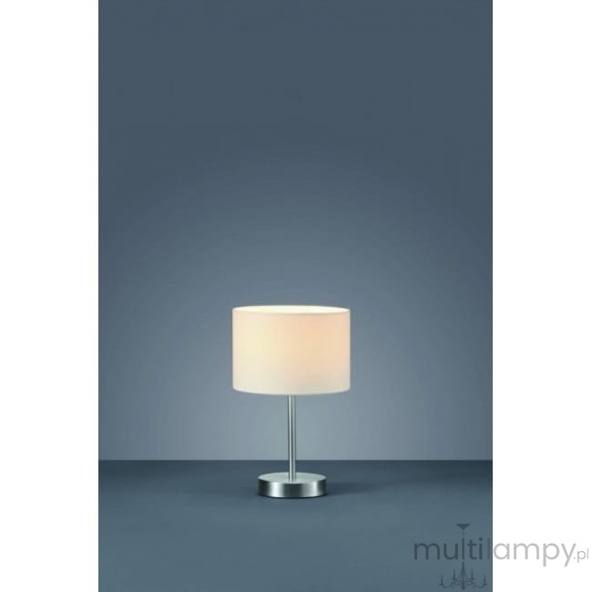 Lampa stołowa 1-punktowa w kolorze białym z okrągłym abażurem i metalową podstawą. Ujęcie frontalne, lampa stoi na gładkiej powierzchni, na tle ciemnoniebieskiej ściany.