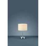 Lampa stołowa 1-punktowa w kolorze białym z okrągłym abażurem i metalową podstawą. Ujęcie frontalne, lampa stoi na gładkiej powierzchni, na tle ciemnoniebieskiej ściany.