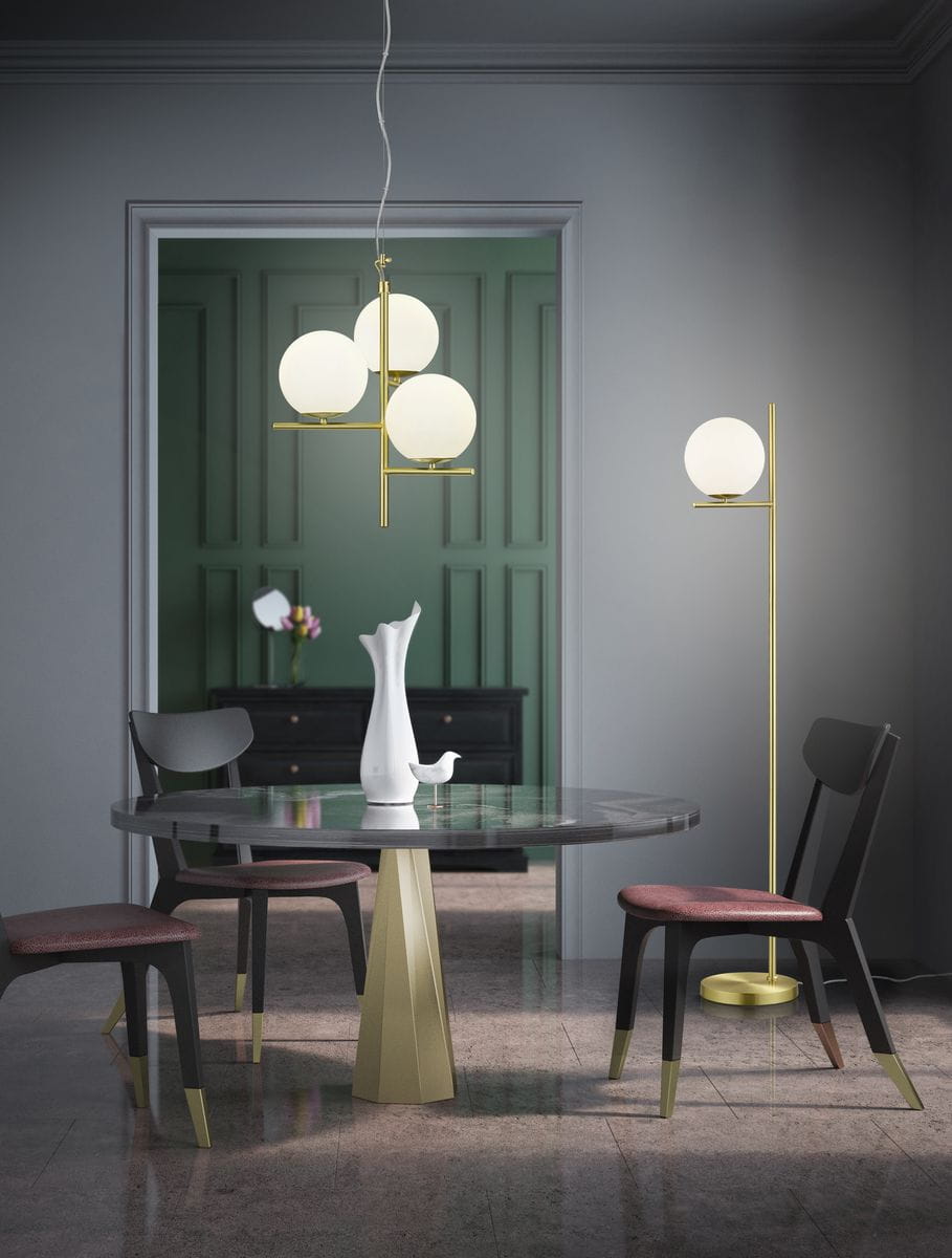 Lampa wisząca Pure 3-punktowa w kolorze mosiądzu z trzema okrągłymi kloszami z białego szkła. Zawieszona na długim przewodzie, umieszczona nad okrągłym stołem w nowoczesnym wnętrzu. W tle widoczny mebel z ciemnego drewna.
