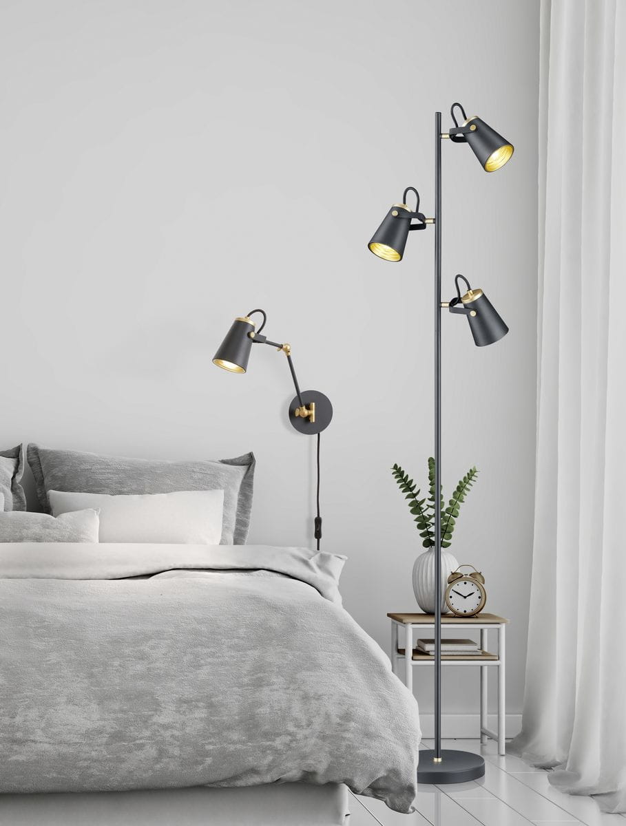 Lampa podłogowa Edward 3-punktowa w kolorze czarnym z metalowymi kloszami, umieszczona obok łóżka. Trzy ruchome źródła światła na smukłym stelażu, w tle widoczna szara pościel i stolik nocny z zegarem oraz rośliną.
