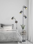Lampa podłogowa Edward 3-punktowa w kolorze czarnym z metalowymi kloszami, umieszczona obok łóżka. Trzy ruchome źródła światła na smukłym stelażu, w tle widoczna szara pościel i stolik nocny z zegarem oraz rośliną.