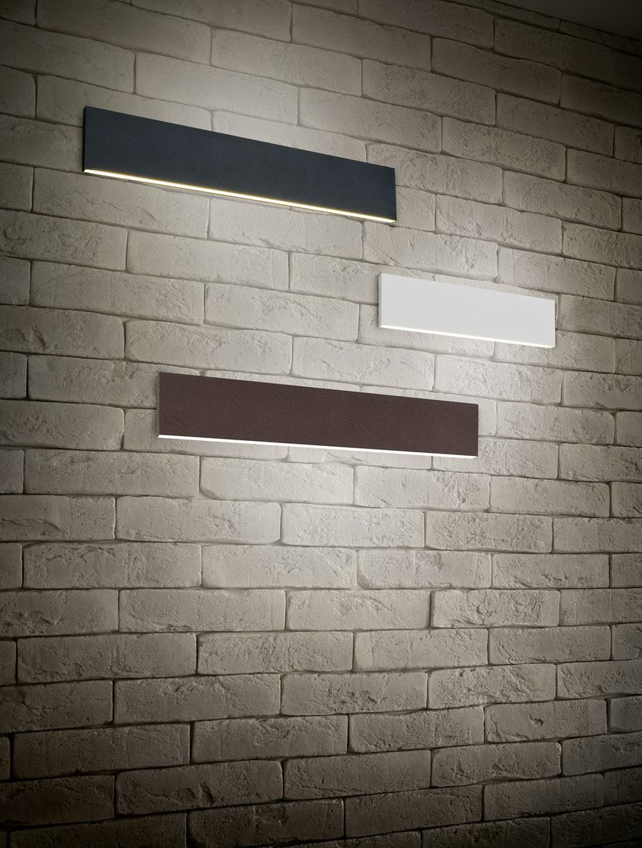 Kinkiet LED Concha 2-punktowy w kolorze antracytowym, zamontowany na ścianie z cegły. Prosta, prostokątna forma z delikatnym podświetleniem, obok białego i brązowego wariantu. Idealny do nowoczesnych wnętrz.