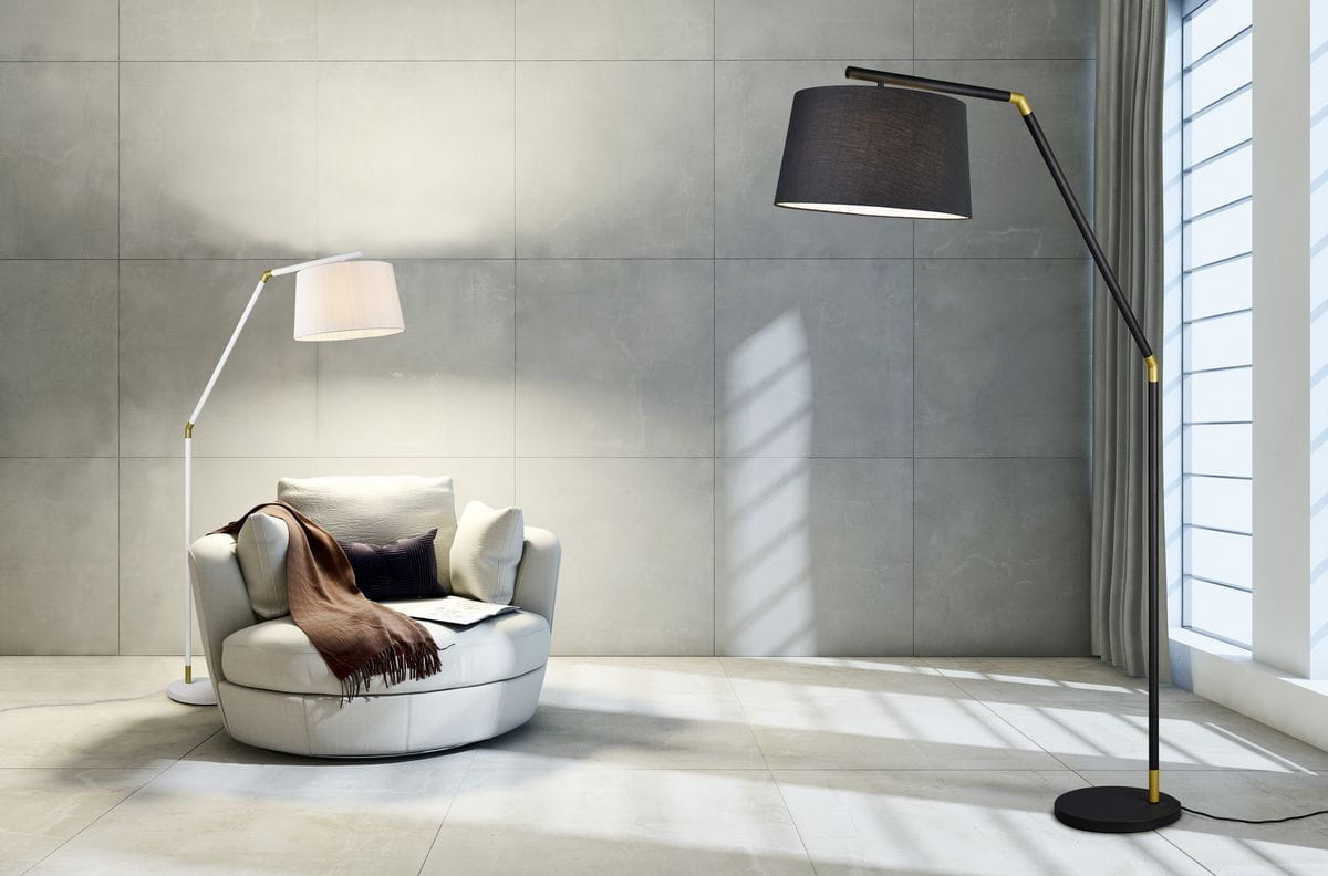 Czarna lampa podłogowa Tracy 1-punktowa, z metalowym stelażem, lekko zakrzywioną nogą i cylindrycznym kloszem. Lampa znajduje się obok wygodnego fotela, na tle jasnej ściany z delikatnymi cieniami.