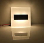 Lumi White LED oprawa schodowa 230V, prostokątna, z mlecznym kloszem, emituje ciepłe światło 3000K. Widoczny czarny element w centralnej części. Ujęcie frontalne, oprawa leży na gładkiej powierzchni, tworząc refleksję.
