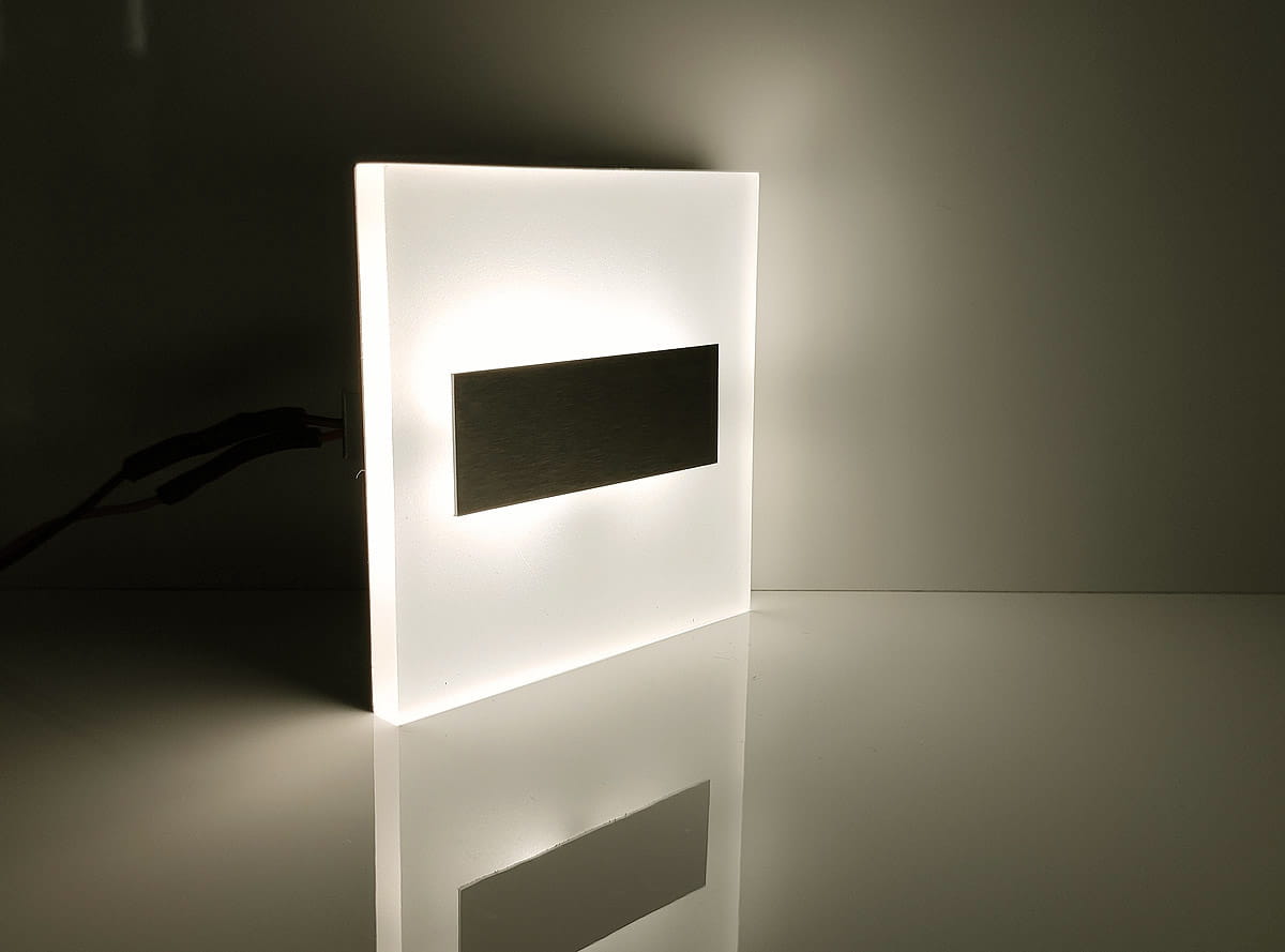 Lumi Black LED oprawa schodowa 230V w barwie neutralnej 4000K, o prostokątnym kształcie. Posiada matową, białą obudowę z metalowym detalem. Ujęcie z boku, produkt leży na gładkiej powierzchni, rzucając delikatny cień.