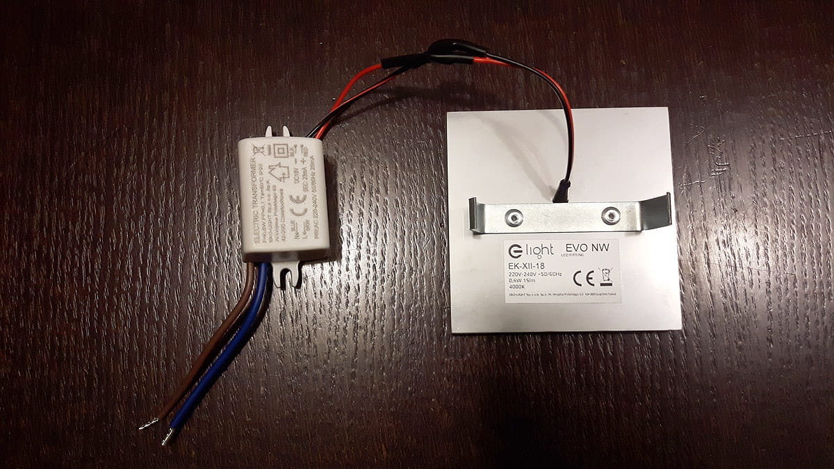 Oprawa schodowa Quadro Black LED 230V z ciepłą barwą światła 3000K. Widoczny kwadratowy panel z metalowym mocowaniem oraz zasilacz, podłączony przewodami w kolorze brązowym, niebieskim i czarnym. Tło ciemne, drewno.