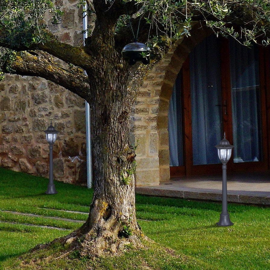 Czarna lampa ogrodowa Vasco H115, stojąca, z metalowym słupkiem i szklanym kloszem, umieszczona w zielonym ogrodzie obok drzewa, na tle kamiennej ściany i okna z zasłonami.