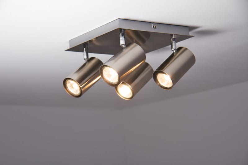 Lampa sufitowa Marley 4-punktowa w kolorze niklu, z czterema cylindrycznymi reflektorami zamontowanymi na kwadratowej podstawie. Reflektory skierowane w różne strony, emitujące ciepłe światło, zamontowane na białym suficie.