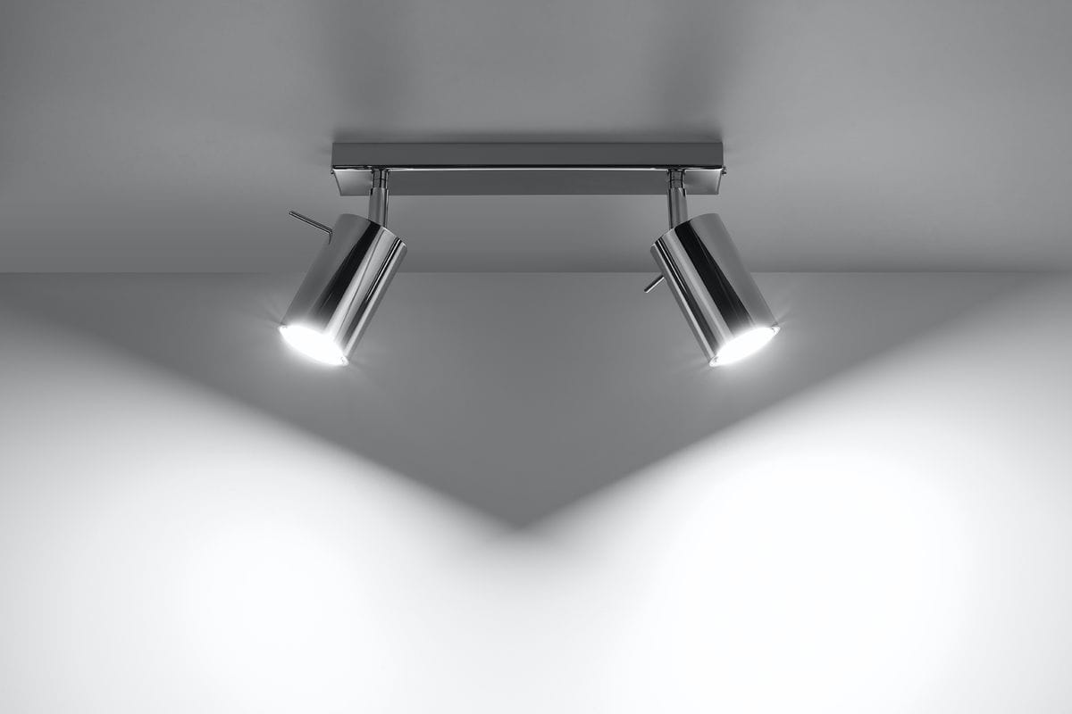 Lampa sufitowa 2-punktowa w kolorze chromowanym z dwoma okrągłymi reflektorami, zamontowana na prostokątnej podstawie. Reflektory skierowane w różne strony, emitują jasne światło, tworząc kontrastowe cienie na gładkiej powierzchni.