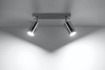 Lampa sufitowa 2-punktowa w kolorze chromowanym z dwoma okrągłymi reflektorami, zamontowana na prostokątnej podstawie. Reflektory skierowane w różne strony, emitują jasne światło, tworząc kontrastowe cienie na gładkiej powierzchni.