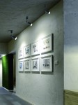 Lampa sufitowa Stick Nero w kolorze białym, 1-punktowa, zamontowana na suficie, skierowana na ścianę z obrazami w białych ramach. Widoczny sufit o teksturze, jasne otoczenie z elementami zieleni w tle.