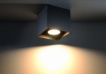 Czarna lampa sufitowa 1-punktowa w kształcie sześcianu, zamontowana na suficie. Emituje ciepłe światło, które oświetla ścianę w tle. Widoczne detale: prostokątne otwory na boku oraz gładka, matowa powierzchnia.