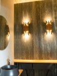Organic LED kinkiet 8-punktowy w kolorze złotym, zamontowany na ciemnej, teksturowanej ścianie. Oświetlenie emituje ciepłe światło, podkreślając nowoczesny design. W tle widoczna okrągła lustro i drewniany stół z czarną umywalką.