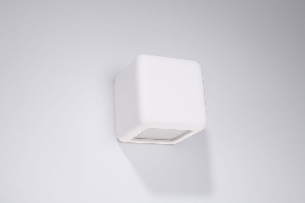 Kinkiet Nesta 1-punktowy, ceramiczny, w kształcie sześcianu, w kolorze białym. Montowany na ścianie, z widoczną przestrzenią na żarówkę w środku. Minimalistyczny design na jednolitym tle.