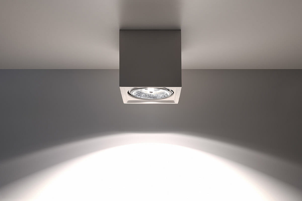Lampa sufitowa Seida SL.0850 w kształcie sześcianu, ceramiczna, w kolorze szarym. Zainstalowana na suficie, z widoczną żarówką w środku, emitującą światło w kształcie stożka na ścianę. Prosty, nowoczesny design, idealna do oświetlenia wnętrz.
