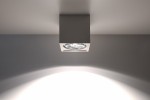 Lampa sufitowa Seida SL.0850 w kształcie sześcianu, ceramiczna, w kolorze szarym. Zainstalowana na suficie, z widoczną żarówką w środku, emitującą światło w kształcie stożka na ścianę. Prosty, nowoczesny design, idealna do oświetlenia wnętrz.
