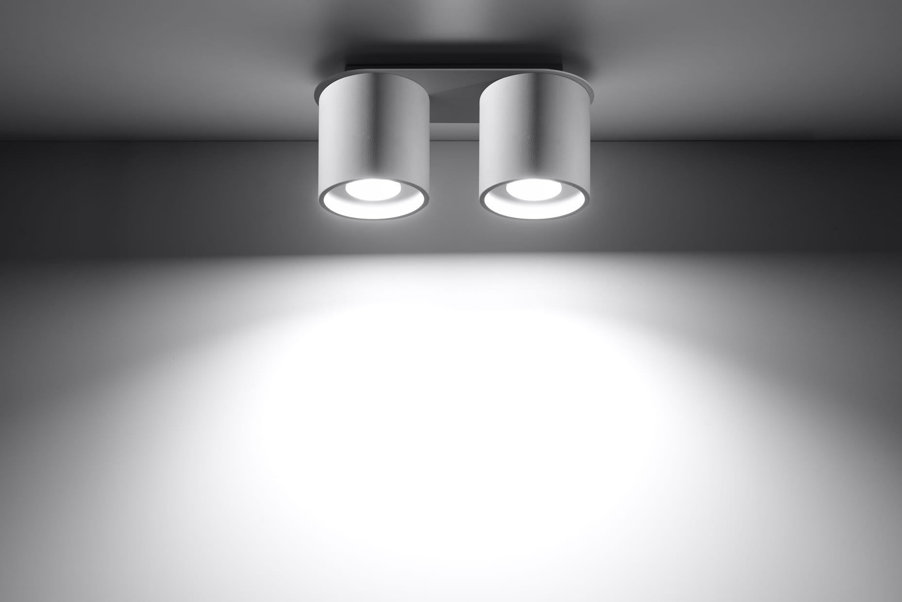 Lampa sufitowa Orbis 2-punktowa w kolorze białym, z dwoma cylindrycznymi kloszami zamocowanymi na płaskiej podstawie. Ujęcie frontalne, pokazujące źródła światła, na jednolitym tle, z wyraźnym oświetleniem.