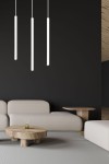 Lampa wisząca Selter 3-punktowa w kolorze białym, składająca się z trzech długich, cylindrycznych elementów zawieszonych na cienkich linkach. Produkt umieszczony w nowoczesnym wnętrzu z ciemną ścianą, obok jasnych mebli i stolika.