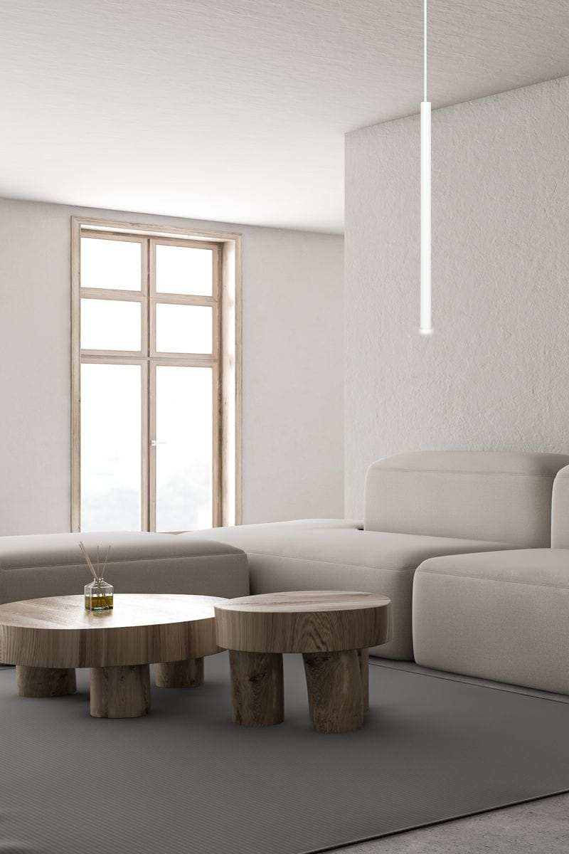 Biała lampa wisząca Selter 553/1, wąska i prosta, zawieszona na długim przewodzie. Produkt widoczny w nowoczesnym wnętrzu z jasnymi ścianami i dużym oknem. W tle szara podłoga oraz drewniane stoliki kawowe.