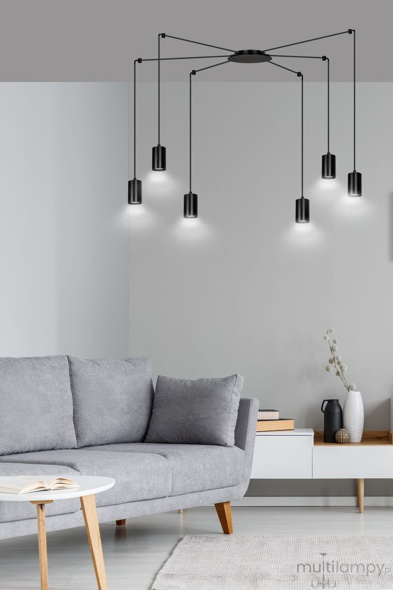 Lampa wisząca Traker 6-punktowa w kolorze czarnym, z sześcioma cylindrycznymi kloszami zawieszonymi na cienkich linkach. Ujęcie frontalne, lampa umieszczona w nowoczesnym wnętrzu z szaro-beżową kanapą i minimalistycznym stolikiem.