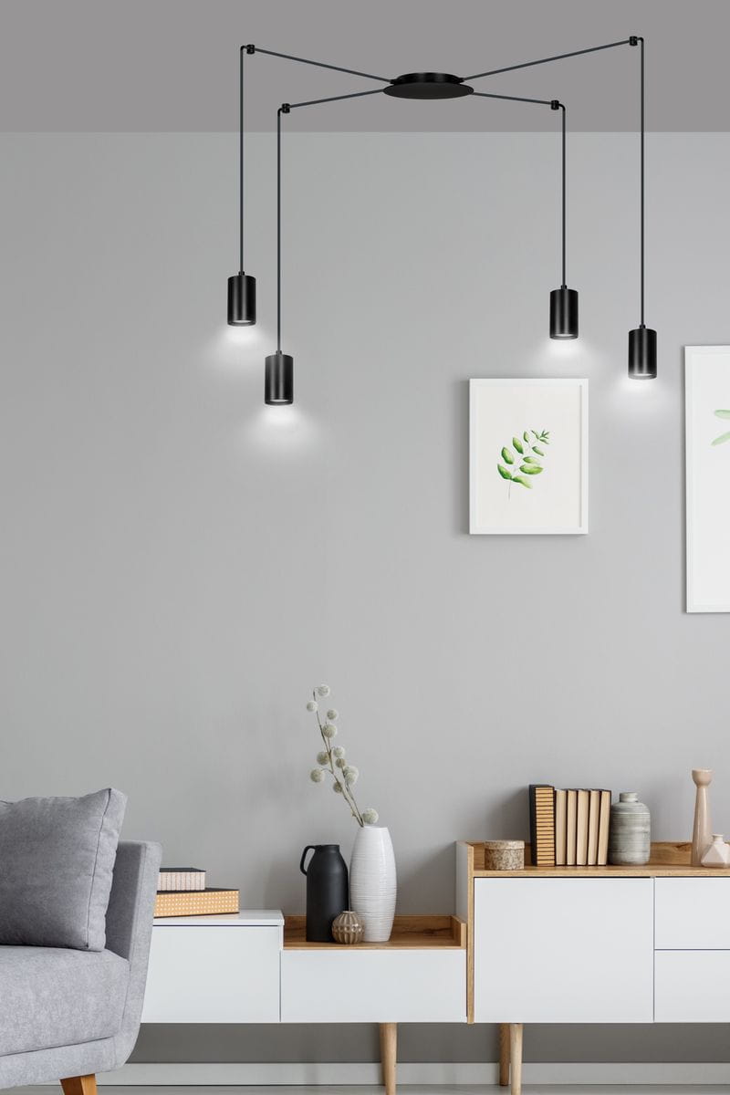 Czarna lampa wisząca Traker 4-punktowa z czterema cylindrycznymi kloszami zawieszonymi na cienkich linkach. Ujęcie z boku, lampa montowana do sufitu, w tle minimalistyczne wnętrze z szaro-białymi meblami i roślinami w doniczkach.