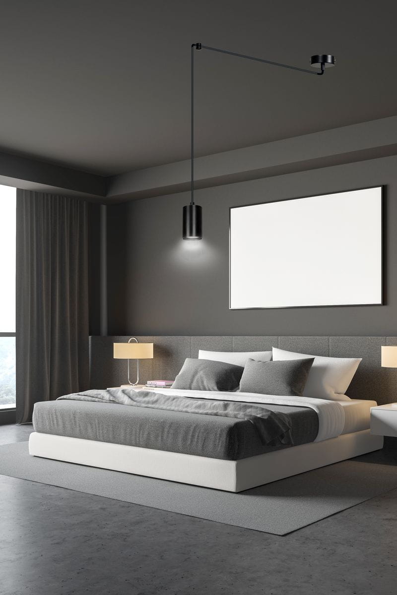 Czarna lampa wisząca 1-punktowa Traker, umieszczona nad łóżkiem, z długim kabelkiem. Wnętrze w nowoczesnym stylu, z szarymi ścianami i białym łóżkiem. W tle widoczne okno z zasłonami oraz lampka nocna obok łóżka.