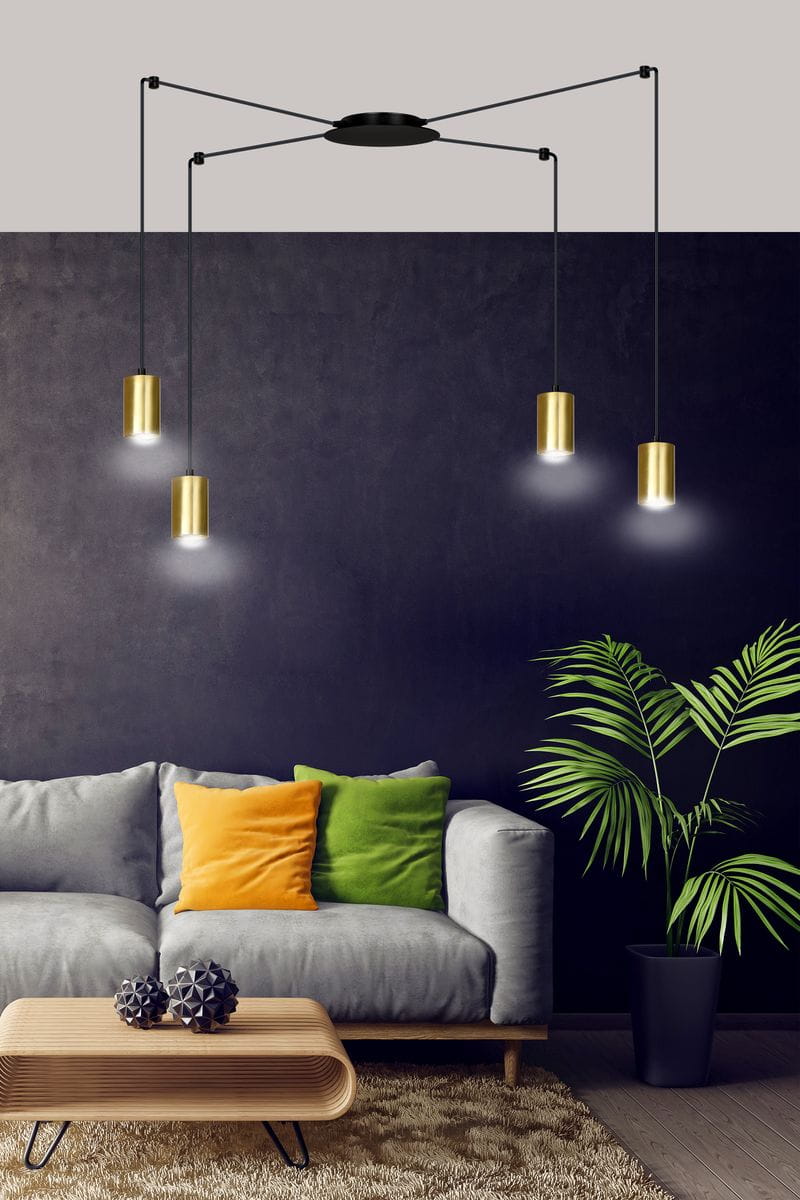 Lampa wisząca Traker 4-punktowa z czarnym stelażem i złotymi kloszami, zawieszona na długich linkach. Klosze mają cylindryczny kształt, emitując ciepłe światło. W tle widoczna szara kanapa z kolorowymi poduszkami oraz roślina w doniczce.