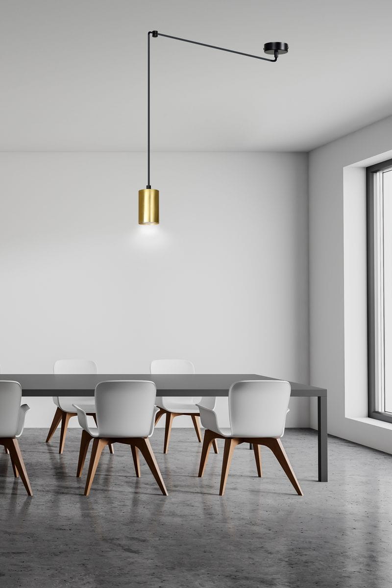 Lampa wisząca Traker 1-punktowa w kolorze czarnym i złotym, zawieszona na długim, czarnym przewodzie. Cylindryczny klosz ze złotego metalu, emitujący światło, umiejscowiony nad prostokątnym stołem w minimalistycznym wnętrzu z białymi krzesłami.