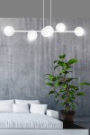 Lampa wisząca Rossi 6-punktowa w białym kolorze z transparentnymi kloszami. Zawieszona na trzech linkach, umieszczona nad jasnoszarym sofą. Tło to surowa, szara ściana, w pobliżu roślina w doniczce.