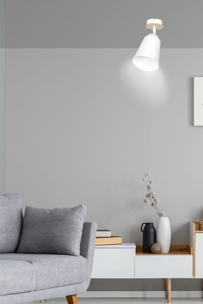 Biała lampa sufitowa typu spot o stożkowej formie, zamontowana na suficie. Wnętrze klosza odsłania żarówkę. W tle szary mural, szare sofa z poduszkami oraz nowoczesny stolik z dekoracjami.
